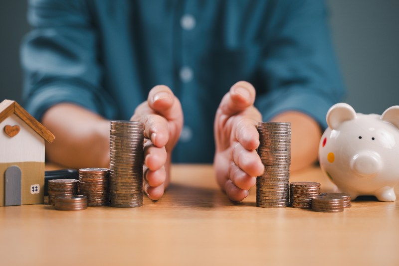 Hands making separate piles of coins by a wooden house and piggy bank by the Law Offices of Robert E. O’Connor